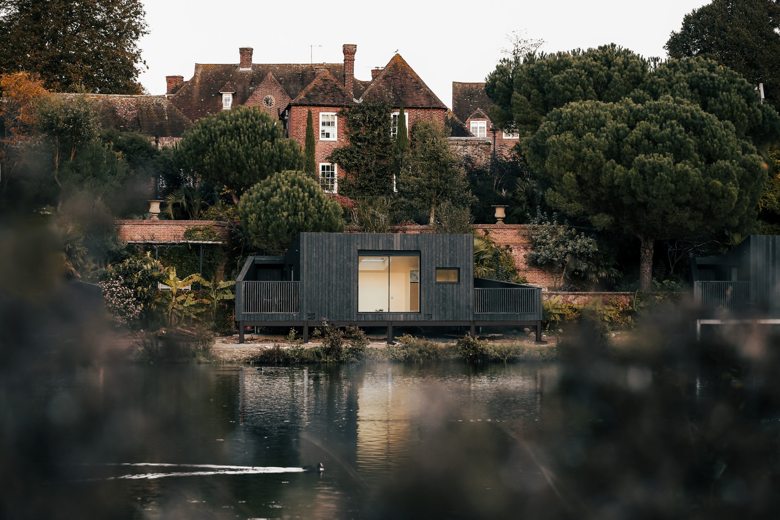 A modern dark cabin with large windows sits beside a calm body of water, reflecting its structure and the surrounding greenery. In the background, a traditional brick house with chimneys and gabled roofs is nestled among tall trees, creating a serene and picturesque landscape.