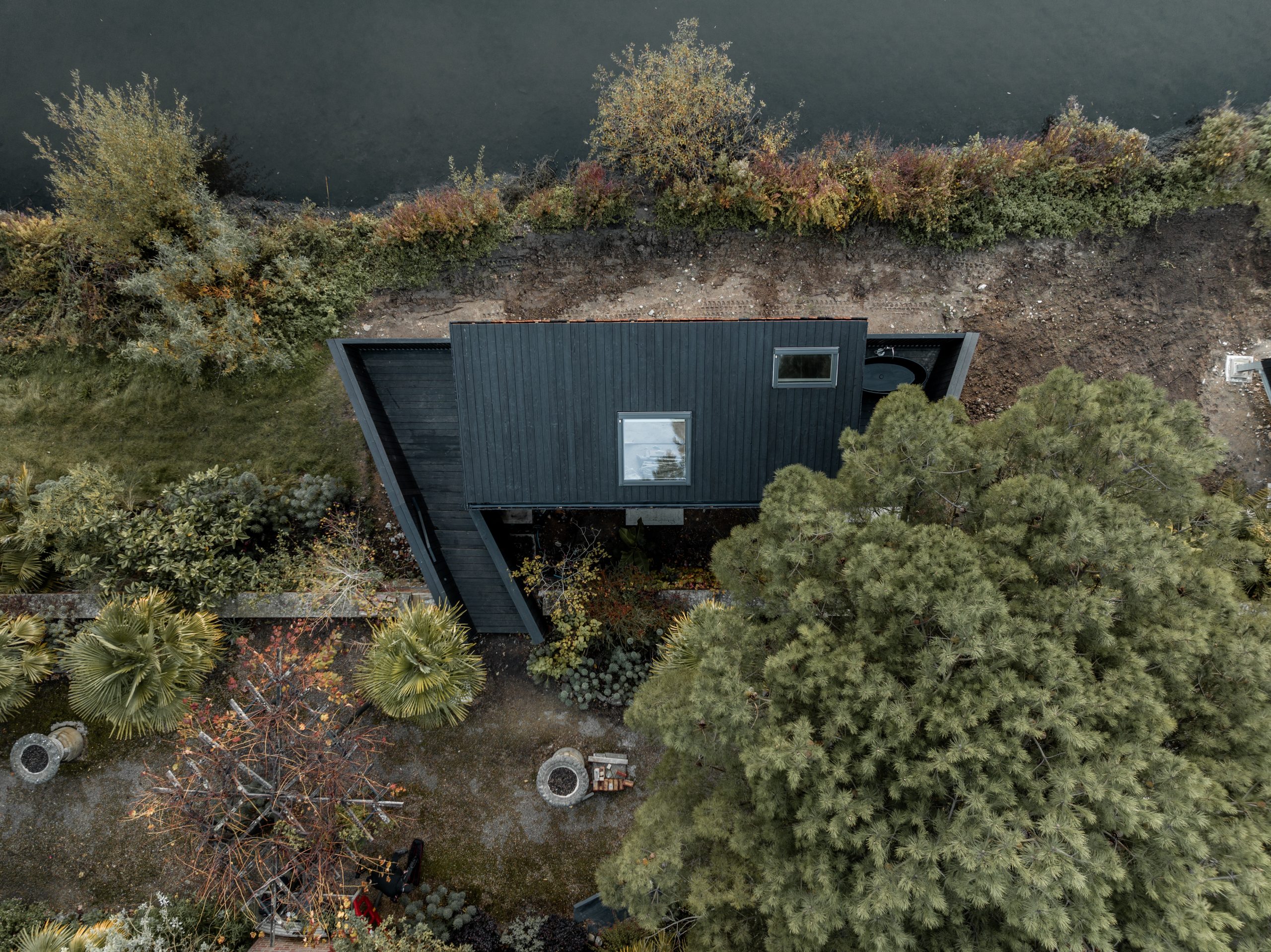 Aerial view of a modern black house with a flat roof and large windows, nestled among lush greenery. The property features landscaped gardens with pathways and circular seating areas, and is situated near a body of water, possibly a river or lake.