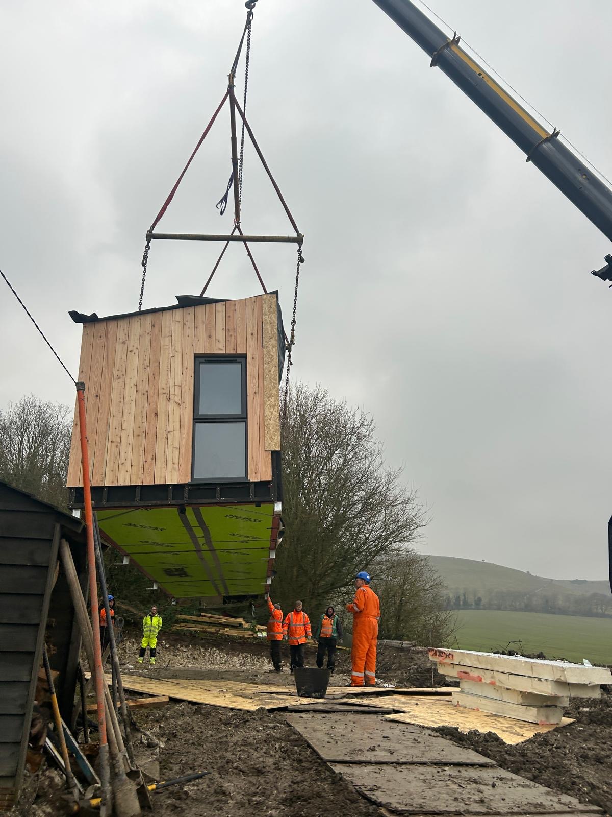 A construction site where a prefabricated building module with wooden paneling and a window is being lifted by a crane. The module is suspended by straps, and several workers in high-visibility clothing and hard hats are guiding its placement on the ground.