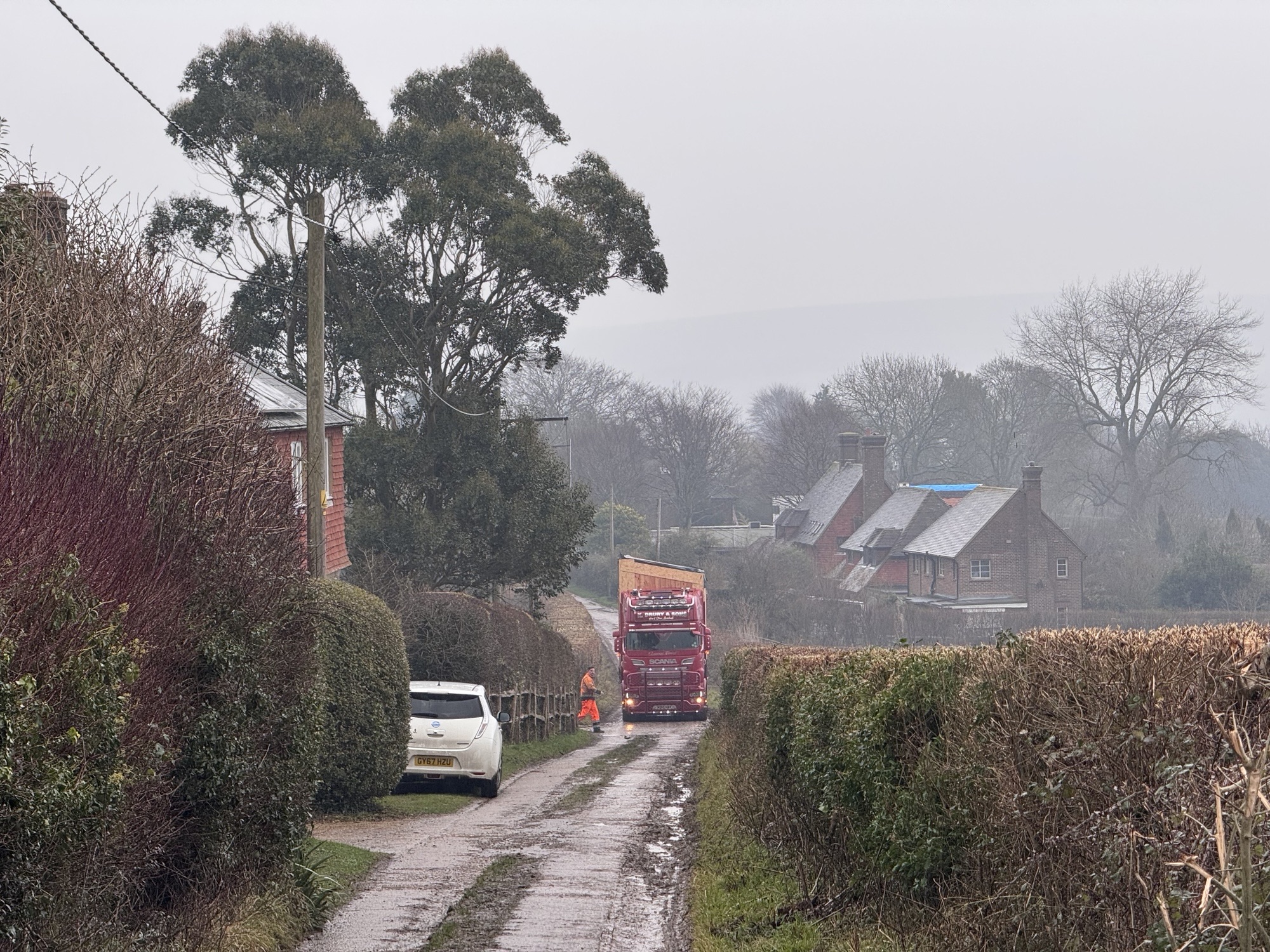 A narrow country road bordered by hedges and trees, with a white car parked on the grass to the left and a large red truck driving down the road. Two people in orange high-visibility clothing stand nearby. Brick houses with sloped roofs are visible in the misty, overcast background.