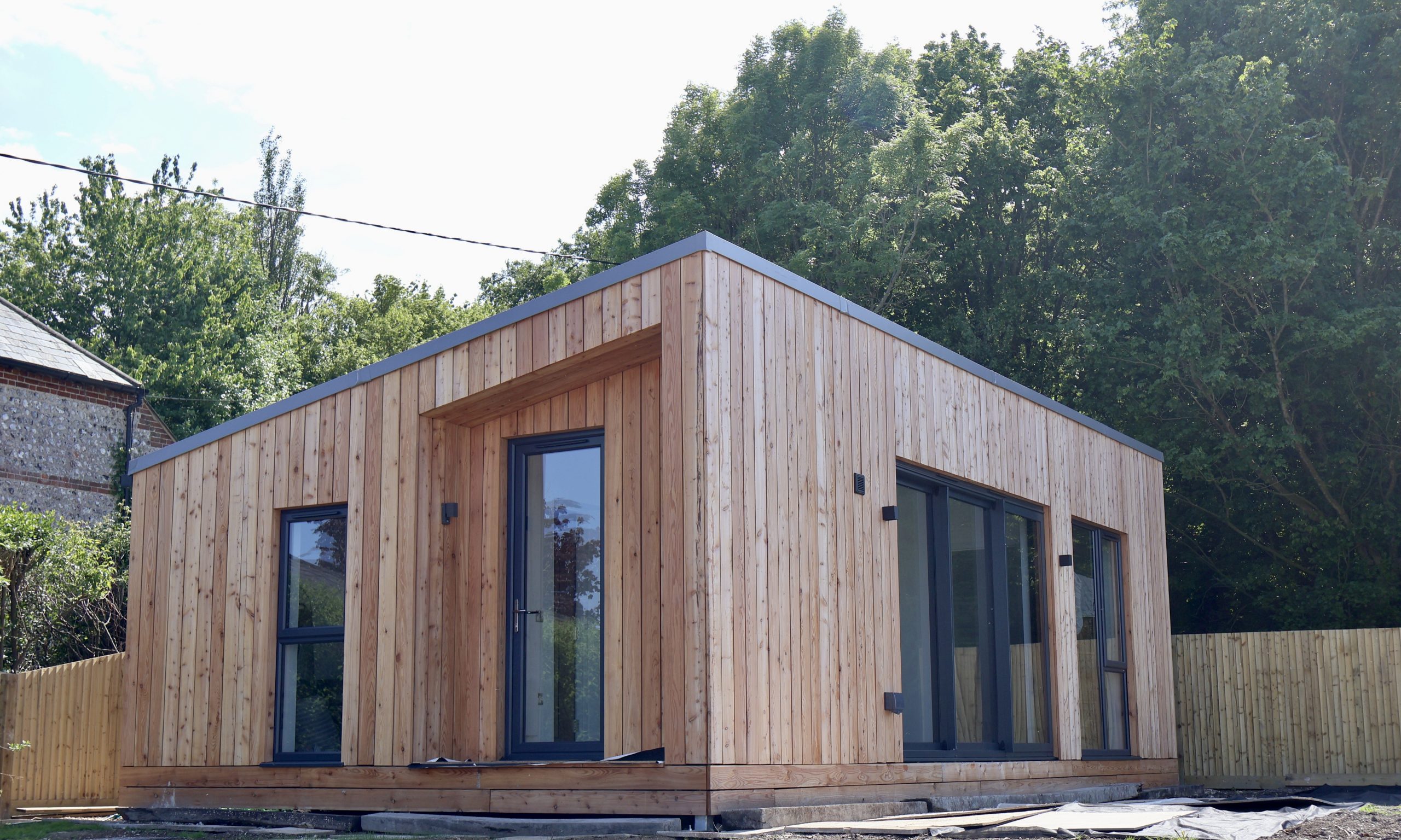 A modern, single-story wooden house with a flat roof and large windows, surrounded by trees and bordered on one side by a wooden fence