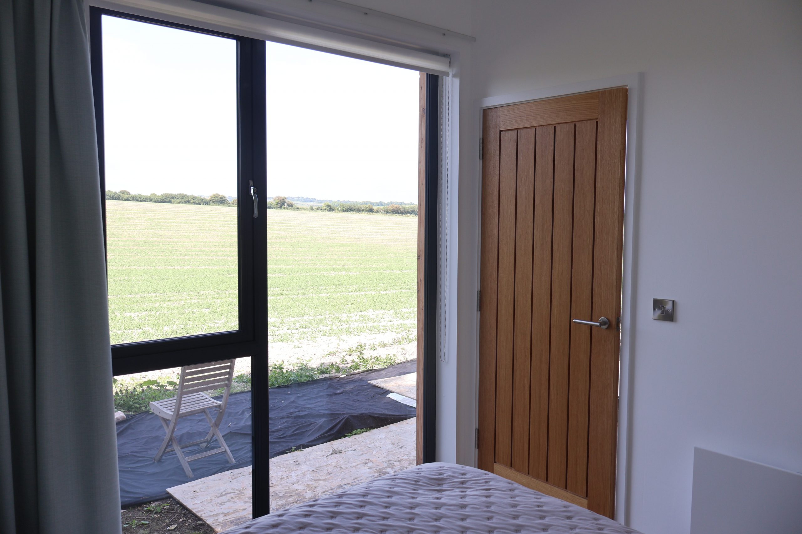 A room with a large window and a wooden door, offering a view of green fields and distant trees. Outside the window, a wooden chair sits on black plastic sheeting. Inside, part of a bed with a quilted blanket is visible in the foreground, and the walls are white.
